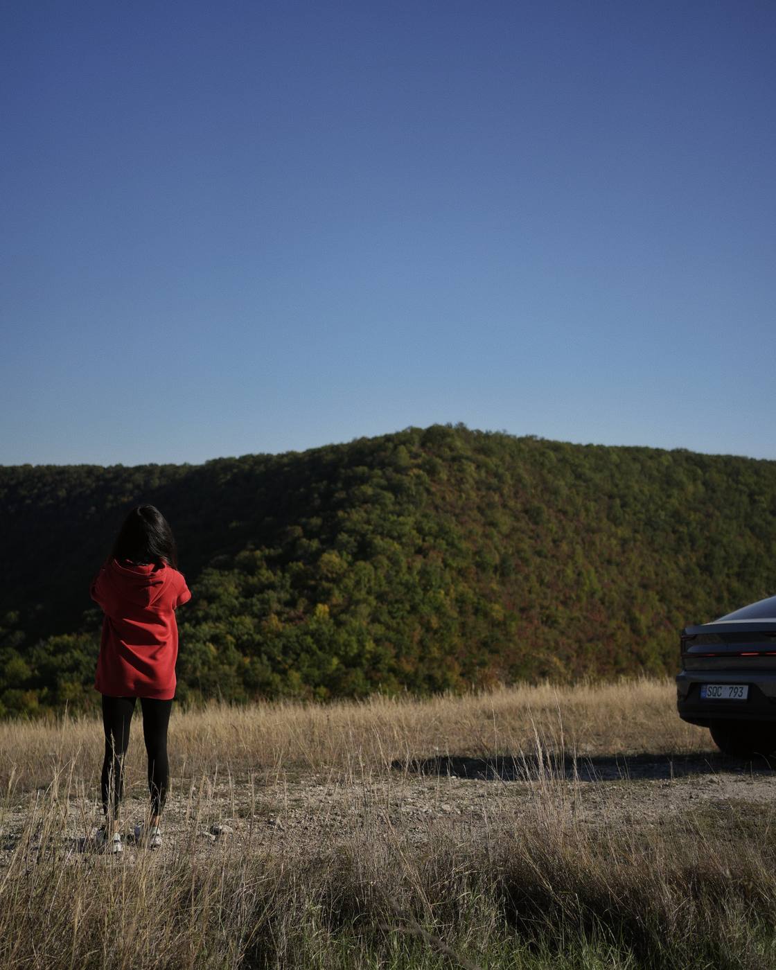 My better half, from behind, in a red jacket, looking at the wooded hill; on the right, the back of the car